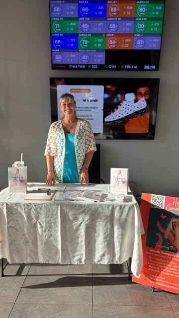 Woman standing at a vendor booth with printed materials and promotional items displayed on a white tablecloth, with digital displays above
