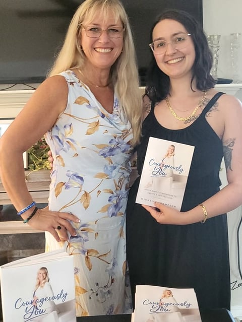 Two women smiling at camera, one wearing a floral dress and glasses, the other in a black tank top holding a book titled Courageously You