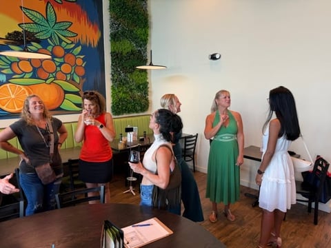 Five women standing in an indoor space with colorful fruit artwork on the wall behind them, appearing to socialize at an event