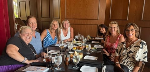 A group of eight adults smiling at a dining table set with food and drinks indoors