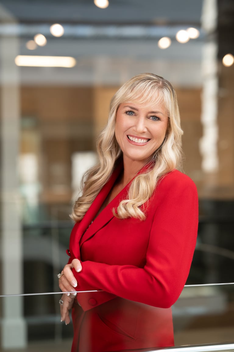 Professional woman with blonde wavy hair wearing a red blazer, smiling at the camera in a modern office setting with blurred background lights.