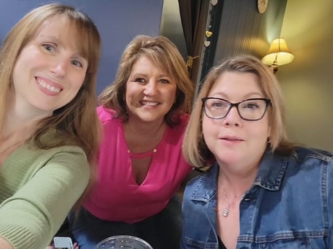 Three smiling women posing together indoors, one wearing a green shirt, one in pink, and one in a denim shirt with glasses