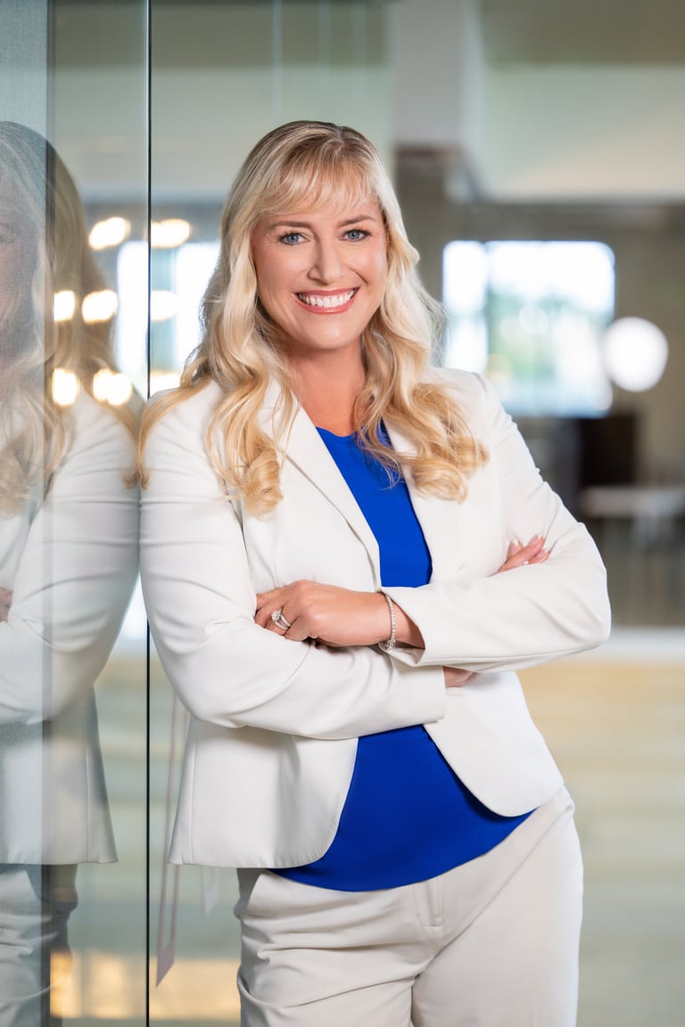 Professional woman with blonde hair wearing a white blazer over a blue top, standing in a modern office with arms crossed