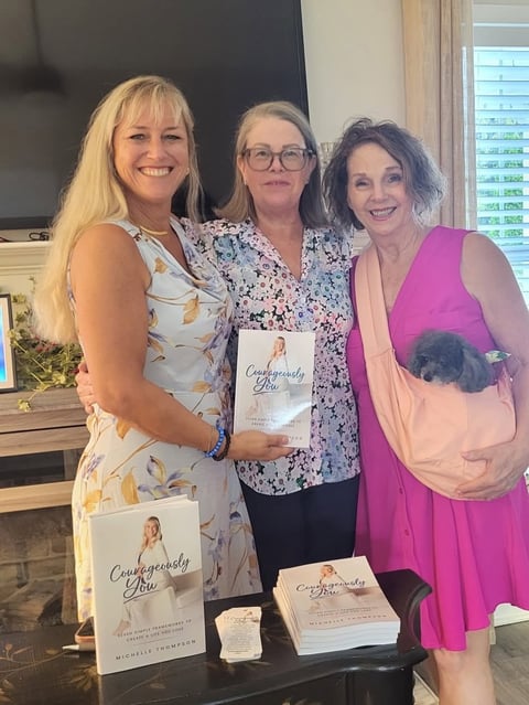 Three women smiling at a book signing event, holding copies of Courageously Brave by Michelle Crawford displayed on table