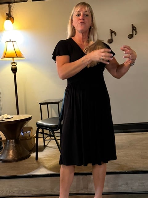 Woman in black dress gesturing expressively while speaking, with musical note decorations on wall behind her