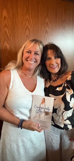 Two smiling women posing together indoors, one in white sleeveless top holding a book, against wooden paneled wall