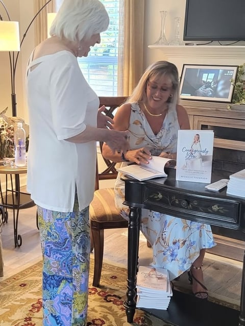 Woman signing a book at a table while another woman in white shirt stands nearby during a book signing event