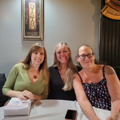 Three women smiling at a table with papers and a phone, indoor setting with framed art on wall behind them
