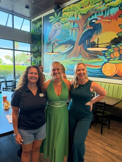 Three smiling women posing together inside a restaurant with colorful nature mural featuring wildlife and fruit on the wall behind them