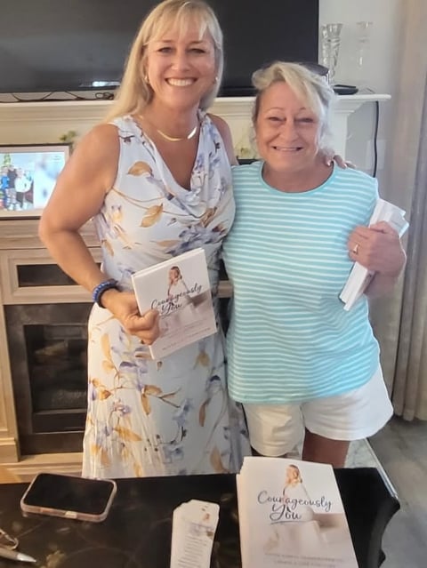 Two women smiling in a home kitchen, holding printed materials, with books on the table in front of them