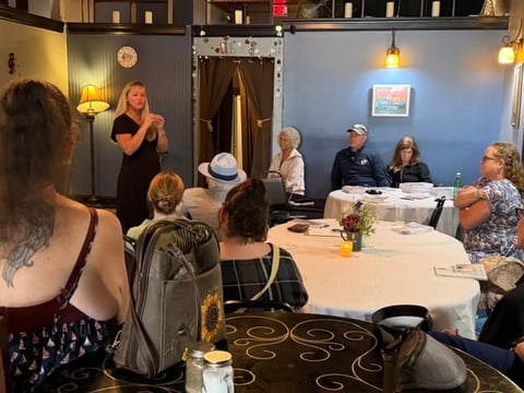 Group of people seated at tables during an indoor event or meeting, with a woman standing and speaking to the audience