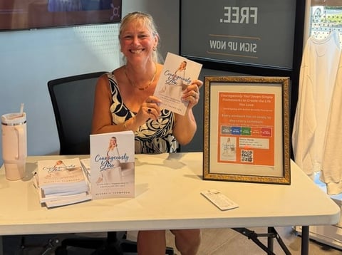 Woman at desk holding up magazine cover, with framed artwork, business cards, and promotional materials displayed