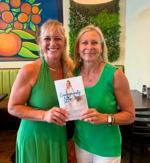 Two women in green shirts holding a book titled Courageously You in an indoor setting with citrus artwork and green plant decor